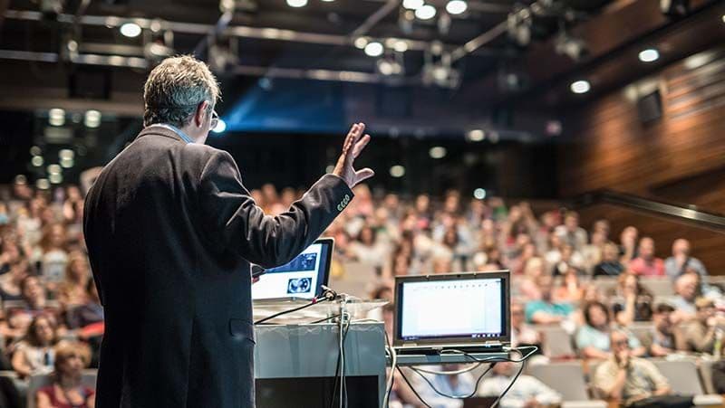 Referent hält eine Präsentation vor einem großen Publikum mit Laptop und Präsentationstechnik